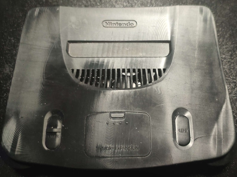 Op de afbeelding: Een zwarte Nintendo 64 console met een geheugenuitbreidingssleuf, een aan/uit-knop en een resetknop. De console heeft een getextureerd oppervlak en een Nintendo-logo aan de bovenkant.