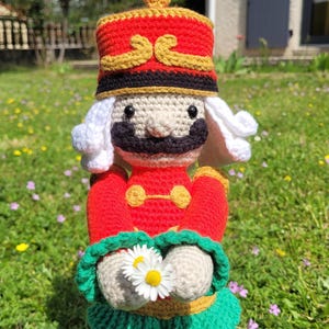 May include: A handmade crochet nutcracker doll. The doll wears a red and gold hat, red jacket, and green skirt. It holds a daisy. The doll has a black mustache and white hair. The doll is standing in a grassy area.