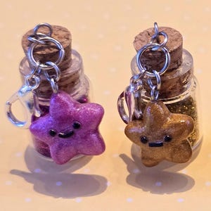 May include: Five small glass bottles with cork stoppers, each filled with glitter and a star-shaped charm. The charms are in various colors: purple, brown, green, blue, and gray. Each charm has a smiling face and is attached to a keychain.