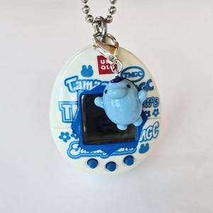 May include: A white Tamagotchi digital pet device featuring blue text and floral graphics, a dark blue screen border, and three blue buttons. A small, light blue manatee charm hangs from a silver lobster clasp attached to a silver ball chain.