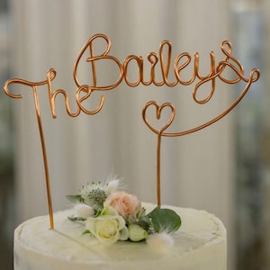 May include: A white wedding cake with a copper wire cake topper that says "The Baileys" with a heart.
