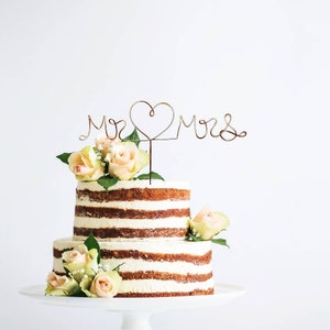 May include: A two-tiered wedding cake decorated with fresh flowers and a wire topper that says "Mr. & Mrs." with a heart in the middle.