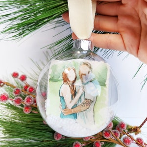 May include: A clear glass Christmas bauble with a watercolour painting of a couple inside. The bauble is filled with white glitter and has a white ribbon attached.