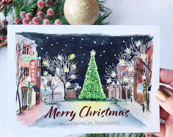 May include: A Christmas card with a watercolor painting of a town square at night, featuring a decorated Christmas tree and the words "Merry Christmas" and "From Franklin, Tennessee". The card is surrounded by Christmas ornaments and greenery.