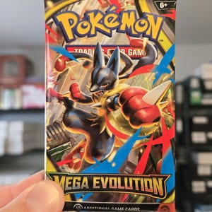 以下が含まれることがあります： ルカリオのダイナミックなイラストが描かれた、未開封のポケモンカードパック。 パックは黒、赤、青を基調とし、「ポケモン」と「メガ進化」の文字が大きく表示されています。 追加のゲームカードが10枚入っています。