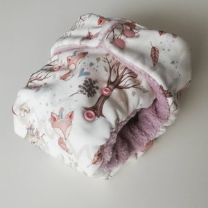 Può includere: Un pannolino di stoffa bianco con una stampa boschiva stravagante con volpi, alberi e foglie nei toni del rosa, marrone e grigio. L'interno è foderato con un materiale morbido color lavanda e si chiude con bottoni a pressione rosa.
