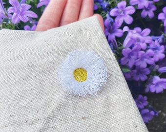 May include: A white canvas tote bag with a hand-stitched white daisy with a yellow centre.