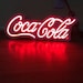 May include: A white neon sign with the word 'Coca-Cola' in red neon lettering. The sign is illuminated and casting a red glow on the surface below.