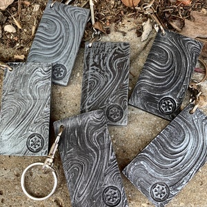 Könnte beinhalten: Sechs silberne Schlüsselanhänger mit einem schwarzen und weißen Marmormuster. Jeder Schlüsselanhänger ist mit einem kleinen silbernen Emblem mit einem Symbol verziert. Die Schlüsselanhänger liegen auf einer braunen Oberfläche.