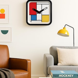 Op de afbeelding: Vierkante wandklok met een zwart frame en een kleurrijk ontwerp in Mondriaanse stijl. De wijzerplaat heeft rode, blauwe, gele en witte blokken. Zwarte wijzers geven de tijd aan. De klok is aan een witte muur bevestigd.