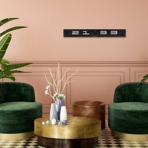 May include: A black flip clock with white numbers displaying the time 2:13. The clock is mounted on a pink wall above a gold coffee table with two green velvet armchairs. The coffee table has a wooden base and a gold top. There are three decorative vases on the table, one blue, one white, and one gray.