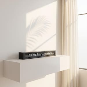 May include: A black digital clock displaying "It's eight oh eight" on a white shelf against a white wall. The clock has a rectangular shape and a modern design. The text is in a clean, sans-serif font. The room has a minimalist aesthetic.
