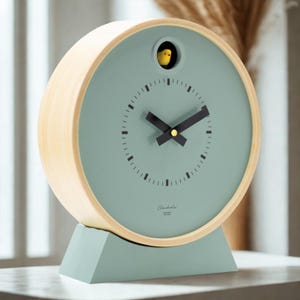 May include: A round, mint green cuckoo clock with a light wood frame and base. The clock face features black hands and a small yellow bird. The clock has the text "Cuculola" on the face.