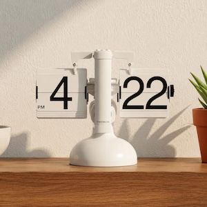 Op de afbeelding: Een witte Twemco flipklok met een centrale standaard en een ronde voet. De klok geeft de tijd 4:22 PM weer met grote zwarte cijfers op witte flip-panelen. De merknaam Twemco staat in kleine zwarte letters op de centrale verticale steun.