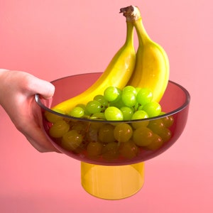May include: A clear glass bowl with a yellow stand, filled with green grapes and two yellow bananas. The bowl is a deep burgundy color.