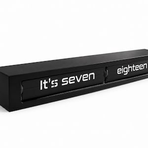 May include: A rectangular black word clock featuring a horizontal display with white text that reads It's seven eighteen. The device has a sleek, matte black finish and a mechanical flip-style mechanism for showing the time in words.