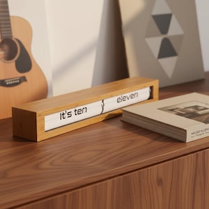 May include: A rectangular wooden desk clock displaying the time as "It's ten" and "eleven". The clock has a white face with black lettering and is set on a wooden surface. A book and artwork are in the background.