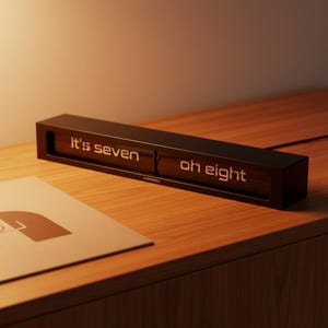 May include: A rectangular, dark brown digital clock displaying "It's seven" and "oh eight" in white illuminated text. The clock sits on a wooden surface, with a matching wooden frame. A piece of paper with a graphic is in the foreground.