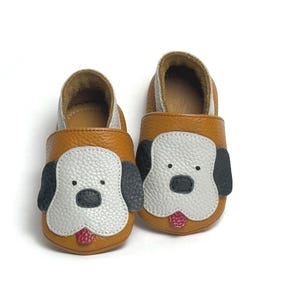 May include: A pair of soft-soled baby shoes in mustard yellow leather, featuring a white dog face appliqué with black ears, black nose, and a small red tongue detail. The shoes have a textured leather finish and a simple slip-on design.