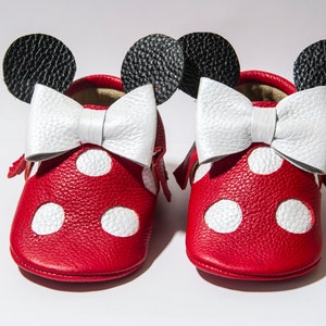 Puede incluir: Zapatos de beb&eacute; de cuero rojo con lunares blancos y un lazo blanco. Los zapatos tienen orejas negras en la parte superior, que se asemejan a un personaje de dibujos animados popular.