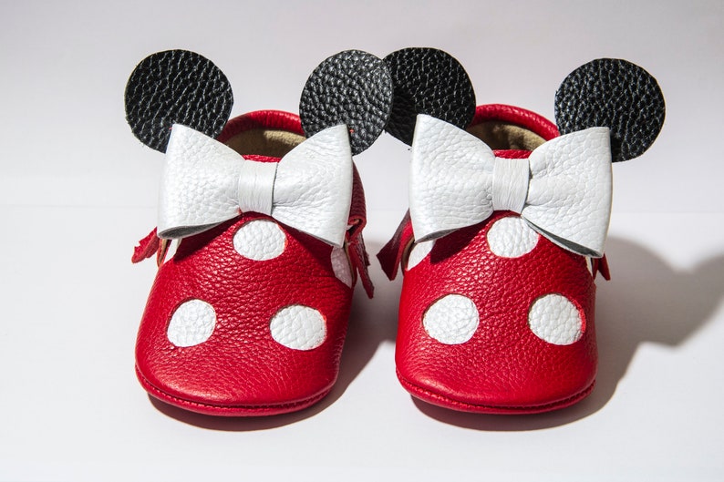 Puede incluir: Zapatos de beb&eacute; de cuero rojo con lunares blancos y un lazo blanco. Los zapatos tienen orejas negras en la parte superior, que se asemejan a un personaje de dibujos animados popular.