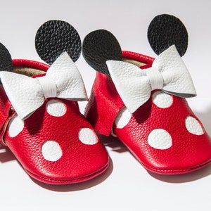 Puede incluir: Zapatos de beb&eacute; de cuero rojo con lunares blancos y un lazo blanco. Los zapatos tienen orejas negras en la parte superior, que se asemejan a un personaje de dibujos animados.