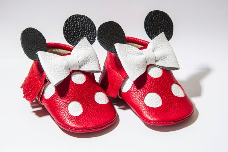 Puede incluir: Zapatos de beb&eacute; de cuero rojo con lunares blancos y un lazo blanco. Los zapatos tienen orejas negras en la parte superior, que se asemejan a un personaje de dibujos animados.