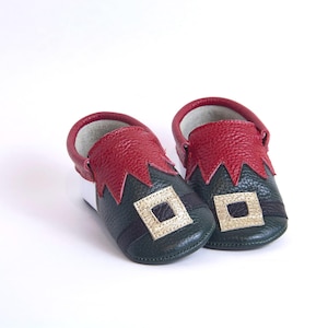 May include: A pair of baby shoes in a festive elf design. The shoes feature a dark green base with a red, jagged-edged collar and a gold square accent. The interior is white, and the shoes are set against a white background.