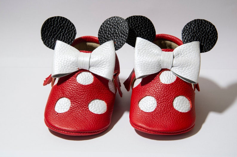 Puede incluir: Zapatos de beb&eacute; de cuero rojo con lunares blancos y un lazo blanco. Los zapatos tienen orejas negras en la parte superior, que se asemejan a un personaje de dibujos animados popular.