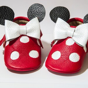 Puede incluir: Zapatos de beb&eacute; de cuero rojo con lunares blancos y un lazo blanco. Los zapatos tienen orejas negras en la parte superior, que se asemejan a un personaje de dibujos animados popular.