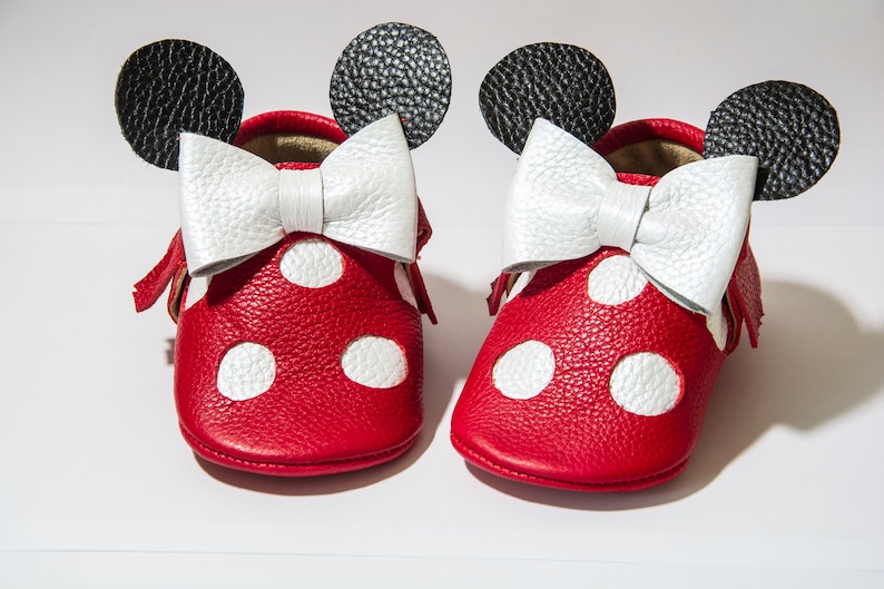 Puede incluir: Zapatos de beb&eacute; de cuero rojo con lunares blancos y un lazo blanco. Los zapatos tienen orejas negras en la parte superior, que se asemejan a un personaje de dibujos animados popular.