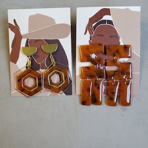 May include: Two pairs of earrings displayed on cards. The first pair is gold with a hexagonal shape and a half-circle design. The second pair is brown and black with a geometric shape.
