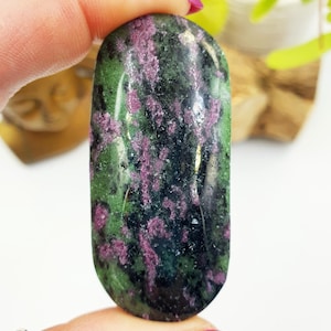 May include: An oval gemstone with a polished surface. The stone displays a mix of green, dark purple, and black colours. The gemstone is held between two fingers, showcasing its natural beauty and unique colour patterns. The stone is approximately 5 cm in length.