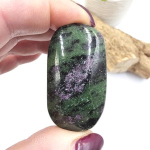 Może przedstawiać: Wypolerowany, owalny kamień szlachetny z mieszanką zielonych, czarnych i fioletowych odcieni. Kamień trzymany jest między palcami, co podkreśla jego gładką, odbijającą światło powierzchnię. Kolory kamienia szlachetnego tworzą uderzający kontrast.