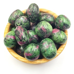 May include: A wooden bowl filled with polished ruby-in-zoisite stones. The stones are oval-shaped, with a mottled green and black appearance, and feature patches of vibrant pink and red. The bowl is made of light-colored wood.