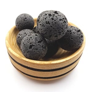 Może przedstawiać: Drewniana miska z czarnymi paskami zawiera kilka ciemnoszarych, porowatych kul z lawy wulkanicznej. Misa jest wykonana z jasnego drewna i ma naturalne usłojenie. Skały lawowe mają szorstką, teksturowaną powierzchnię.