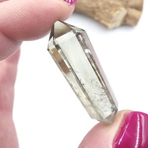 May include: A clear, elongated crystal with a pointed tip. The crystal has a light brown hue and appears to be transparent, with visible internal inclusions. The crystal is held between two fingers, showcasing its polished facets.