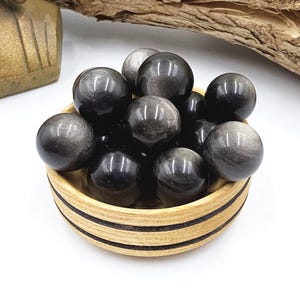May include: A collection of polished, spherical obsidian stones with a metallic sheen, displayed in a small, round wooden bowl. The stones vary in size and reflect light, creating a shimmering effect. The bowl has a natural wood grain finish.