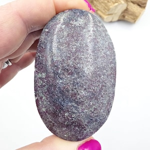 May include: An oval-shaped stone with a textured surface. The stone has a mix of colors, including deep purple, gray, and hints of green. The stone is held in a hand with a pink painted nail.