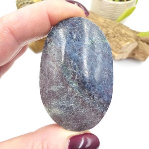 May include: An oval gemstone in shades of blue, purple, and green. The stone is held between fingers, highlighting its polished surface and natural colour variations. The gemstone's texture appears slightly speckled.