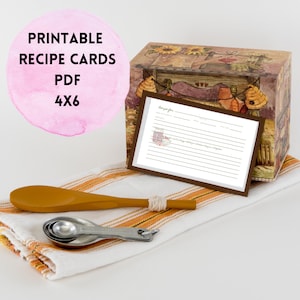 May include: A recipe card box with a decorative design, a wooden spoon, and measuring spoons rest on a striped towel. The box is next to a recipe card with lines for writing. The text "PRINTABLE RECIPE CARDS PDF 4X6" is in a pink circle.