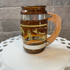 May include: A brown glass mug with a wooden handle. The mug has a white rope design around the middle and images of a penguin, a killer whale, and a seagull. The text "Sea World" is printed on the mug.