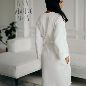 May include: A white, long-sleeved robe with a matching belt and pockets. The text on the image reads: "MODEL IS 5'5" WEARING SIZE S". The robe is made of a textured fabric.