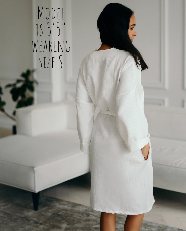 May include: A white, long-sleeved robe with a matching belt and pockets. The text on the image reads: "MODEL IS 5'5" WEARING SIZE S". The robe is made of a textured fabric.