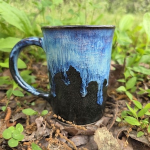 May include: A handmade ceramic mug with a blue and black glaze. The mug has a large handle and a unique drip effect where the blue glaze transitions to black. The mug is sitting on a bed of leaves and small plants.