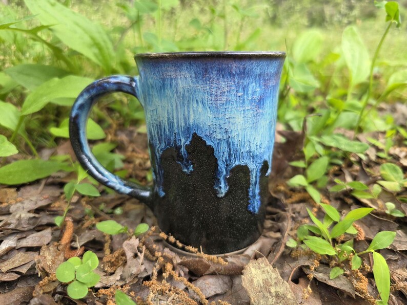 May include: A handmade ceramic mug with a blue and black glaze. The mug has a large handle and a unique drip effect where the blue glaze transitions to black. The mug is sitting on a bed of leaves and small plants.