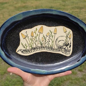 May include: Oval ceramic platter with a dark blue rim and a black speckled interior. The center features a cream-colored design of a skull, mushrooms, and a spine. The mushrooms are yellow and the plants are green.