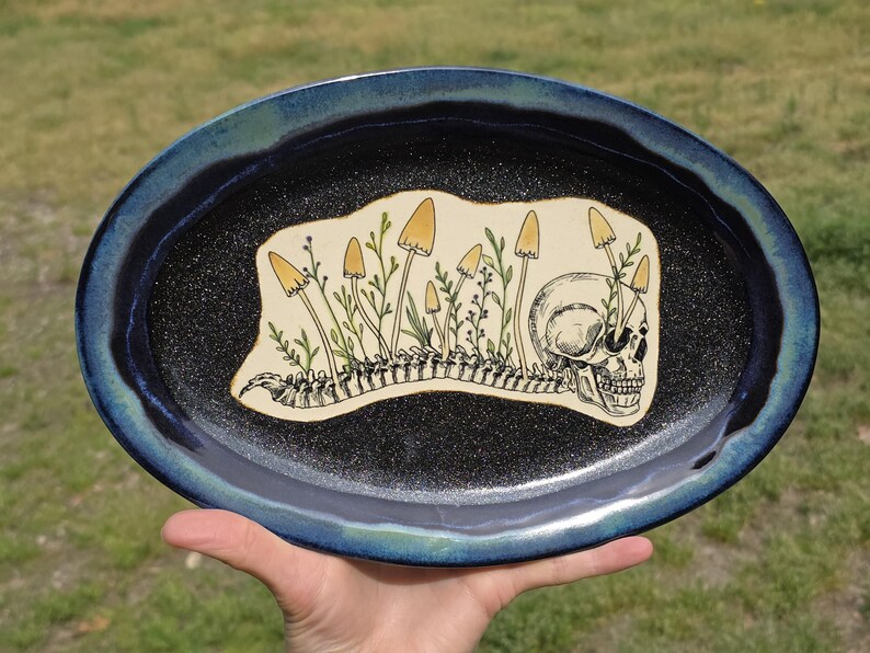 May include: Oval ceramic platter with a dark blue rim and a black speckled interior. The center features a cream-colored design of a skull, mushrooms, and a spine. The mushrooms are yellow and the plants are green.