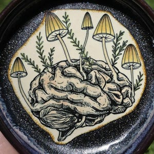 May include: A ceramic trinket dish with a detailed illustration of a brain sprouting mushrooms. The brain is beige and the mushrooms have yellow caps. The dish has a dark brown rim and a sparkly black and blue border.
