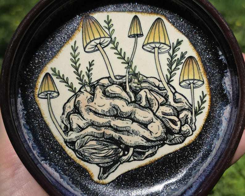 May include: A ceramic trinket dish with a detailed illustration of a brain sprouting mushrooms. The brain is beige and the mushrooms have yellow caps. The dish has a dark brown rim and a sparkly black and blue border.